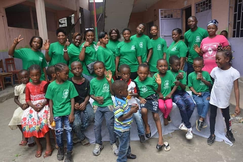 A large group of children and volunteers in matching t-shirts pose together after a Be Real Humanitarian Works distribution